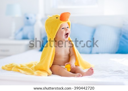 Happy laughing baby wearing yellow hooded duck towel sitting on parents bed after bath or shower. Clean dry child in bedroom. Bathing and washing of little kids. Children hygiene. Textile for infants.