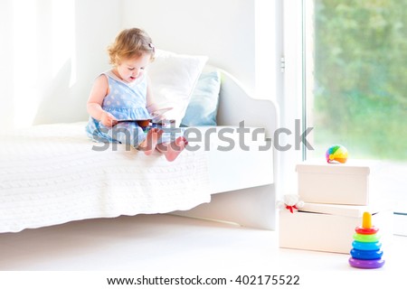 Funny happy toddler girl reading a book and playing with her toy teddy bear in bed. Kids play at home. White nursery. Child in sunny bedroom. Children read and study. Interior for baby and young kid.