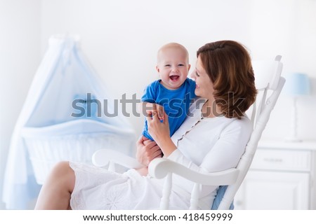 Young mother holding her newborn child. Mom nursing baby. Woman and new born boy in white bedroom with rocking chair and blue crib. Nursery interior. Mother playing with laughing kid. Family at home