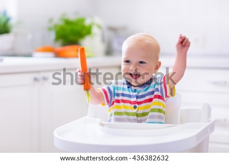 Happy baby sitting in high chair eating carrot in a white kitchen. Healthy nutrition for kids. Bio carrot as first solid food for infant. Children eat vegetables. Little boy biting raw vegetable.