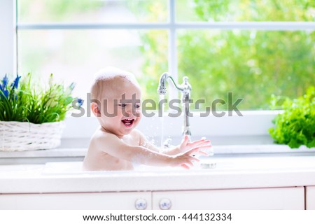 Baby taking bath in kitchen sink. Child playing with foam and soap bubbles in sunny bathroom with window. Little boy bathing. Water fun for kids. Hygiene and skin care for children. Bath room interior