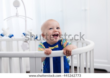 Cute laughing baby standing in a white round bed. White nursery for young children. Little boy learning to stand in his crib. Toys for infant cot. Smiling child playing with toy bear in sunny bedroom.