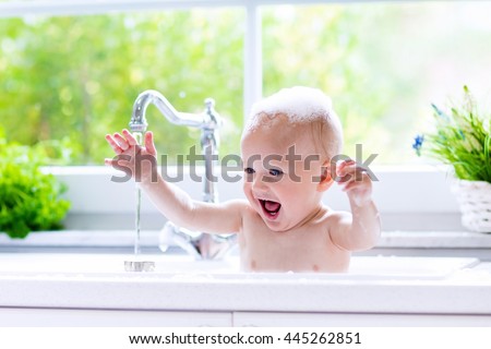Baby taking bath in kitchen sink. Child playing with foam and soap bubbles in sunny bathroom with window. Little boy bathing. Water fun for kids. Hygiene and skin care for children. Bath room interior