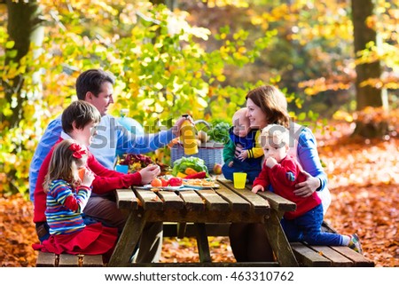 Happy young family with four children grilling meat and making sandwich and salad on a picnic table in sunny autumn park. Barbecue fun for parents with kids on warm fall day. Grill and BBQ party.