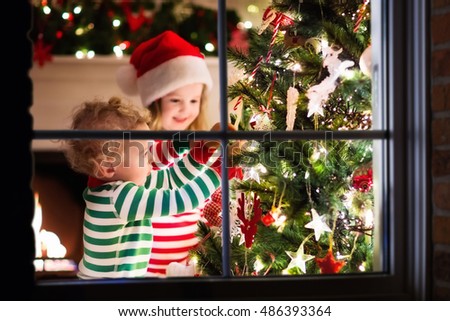 Happy little kids in matching red and green striped pajamas decorate Christmas tree in beautiful living room with traditional fire place. Children opening presents on Xmas eve. View though window.