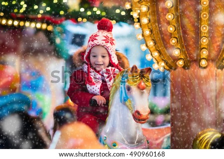 Happy little boy in warm jacket and red knitted Nordic hat and scarf riding carousel horse during family trip to traditional German Christmas market. Kids at Xmas outdoor fair on snowy winter day.
