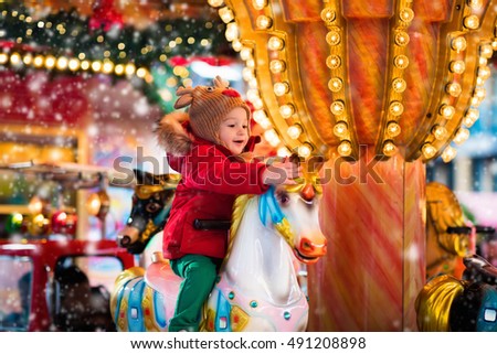 Happy little girl in warm red jacket and knitted reindeer hat riding carousel horse during family trip to traditional German Christmas market. Kids at Xmas outdoor fair on snowy winter day.