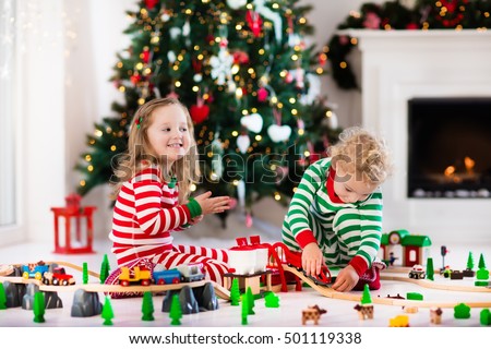 Happy little children in matching pajamas playing with Christmas presents - wooden toy railroad and car. Family Xmas morning in decorated living room with kids gifts, fireplace and Christmas tree.