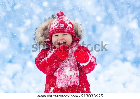 Baby playing with snow in winter. Little toddler boy in red jacket and Xmas reindeer knitted hat catching snowflakes in winter park on Christmas. Kids play in snowy forest. Children catch snow flakes