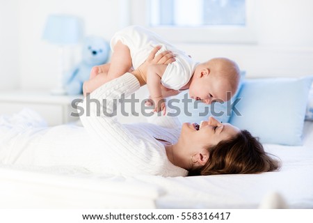Mother and child on a white bed. Mom and baby boy in diaper playing in sunny bedroom. Parent and little kid relaxing at home. Family having fun together. Bedding and textile for infant nursery