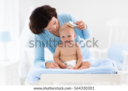 Mother and baby change diaper after bath in white nursery with bed and rocking chair. Little boy on changing table in clean dry nappy. Mom taking care of infant child. Kids room interior and hygiene