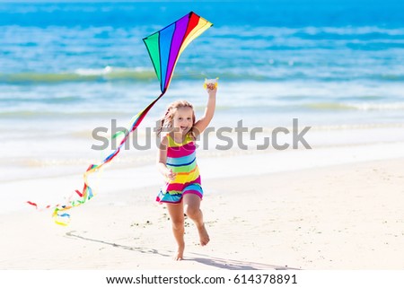 Happy laughing little girl flying a colorful kite running and jumping in sand on beautiful tropical beach during active summer family sea vacation. Kids play on ocean shore. Child with beach toys.
