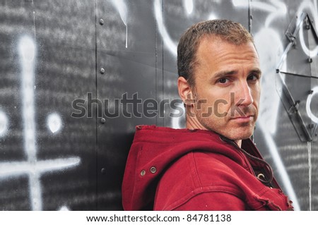 Rugged attractive Caucasian man in red hooded sweatshirt leaning against a graffiti covered wall