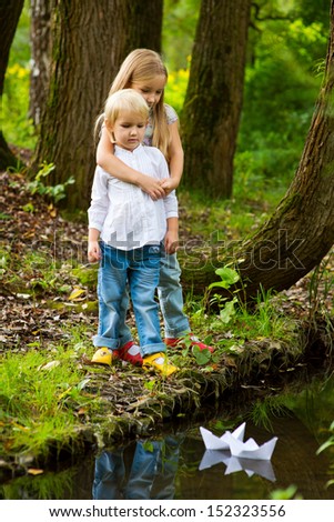 Two adorable european sisters holding each other on scenic river shore and watching on paper boat in river with colorful forest on background