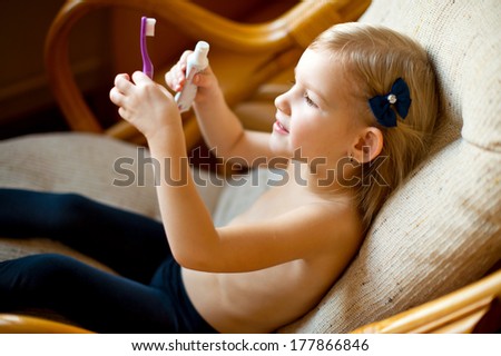 Young happy cute girl with blond hair and bow holding toothbrush and toothpaste in hands and having fun