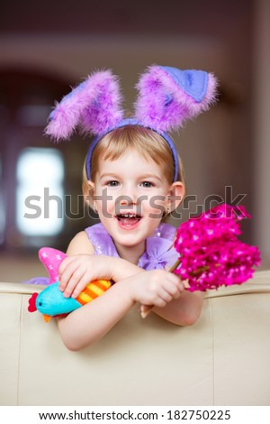 Happy funny little girl with colorful pink flowers and fancy bunny ears on head celebrates easter. Toy han in hands.