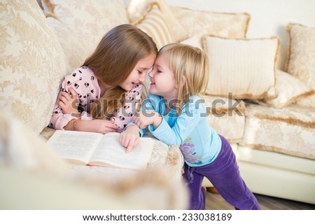 Two cute european sister girls reading a book for children and having fun in room of their home