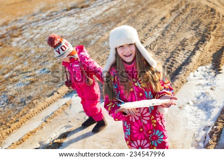 Two sister girls having fun outdoors on winter village road. One girl holding broken piece of ice in hands and smile