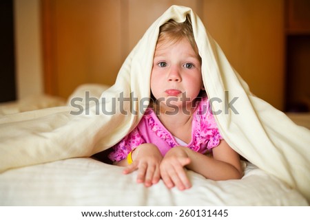 Sad little girl lying on bed under blanket and posing in room of her flat