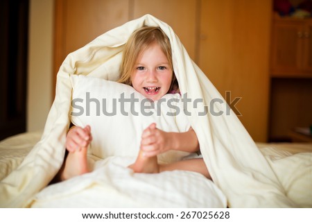 Happy little girl lying on bed under blanket and posing in room of her flat holding a pillow in hands
