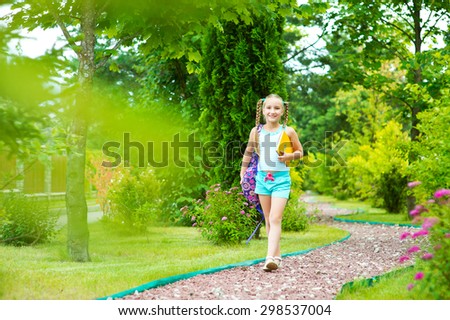 Happy toddler school girl in blue suit walking on road in summer garden with flowers holding book and bag and smiling