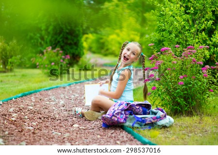 Happy toddler school girl in blue suit sitting on road in summer garden with flowers and reading book with smile