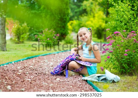 Happy toddler school girl in blue suit sitting on road in summer garden with flowers and put out book from bag