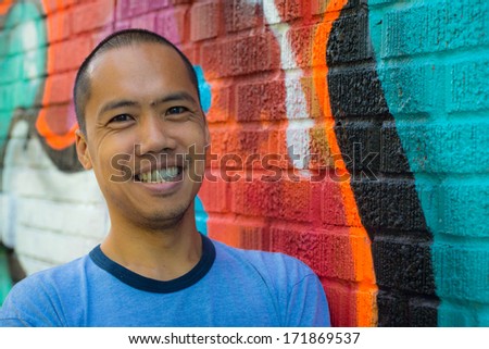 An Asian male stands against colorful graffiti wall in an urban environment, with a big smile