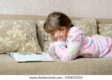 The little girl reads the book lying on a sofa. A house room