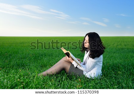 The girl in a business suit reads the book. Sitting on a green meadow
