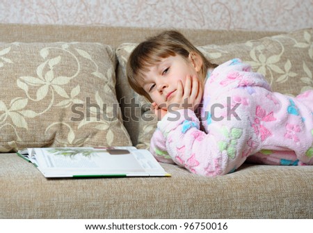 The little girl reads the book lying on a sofa. A house room
