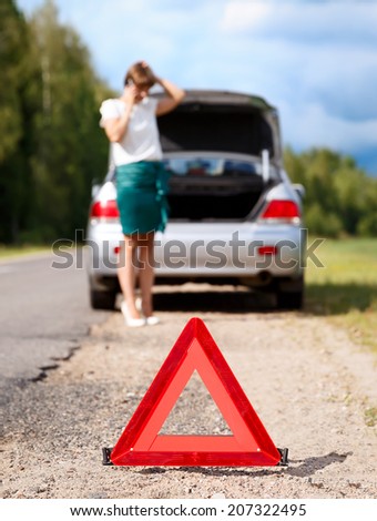 Close up on triangle warning sign with woman calling for assistance after breaking down with her car on background