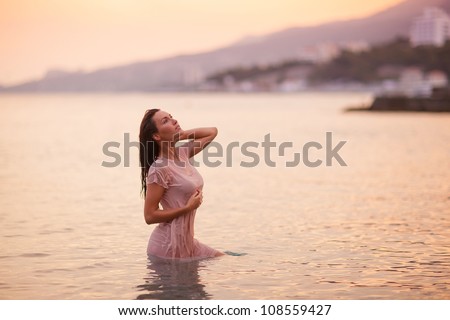 Sensual brunette woman in wet dress in the water at beautiful pink sunset
