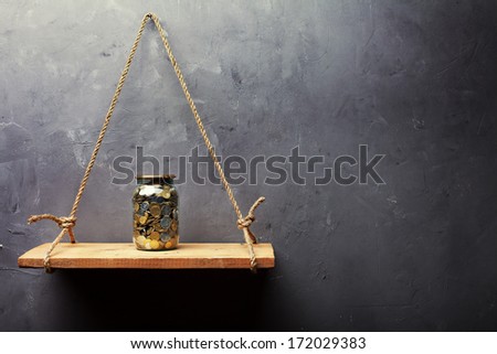 Glass jar with coins on the old wood shelf hanging on rope on textured wall background with copy space