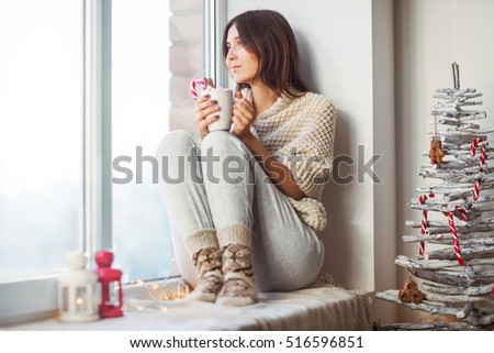 Happy beautiful woman drinking hot coffee sitting on window sill in christmas decorated home. Holiday concept