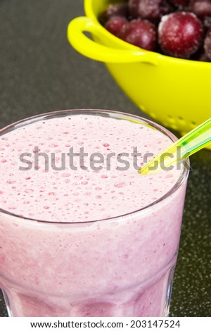 Cherry milk shake in a glass cup with a spoon and a light green colander with frozen cherries on a dark background