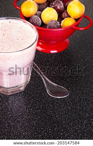 Cherry milk shake in a glass cup with a spoon and a red colander with frozen cherries and cherry-plum on a dark background