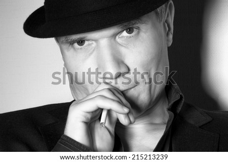 Man in black hat at the age of forty-six years old with a cigarette in his hand looking at the camera on the background of a rough wall with texture