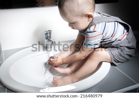Adorable  little boy at the age of two in a striped T-shirt  looking at the flow of water in the sink and washing her hands and feet in the sink