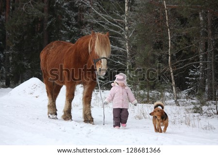 Cute little girl leading big draught horse and small dog by the forest in winter