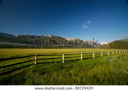 A wooden rail fence stretches across a green meadow hidden in the mountains of northern Wyoming.