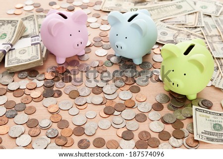 3 Piggy banks, some loose change and dollar bills on a desk.  Main focus on green bank.