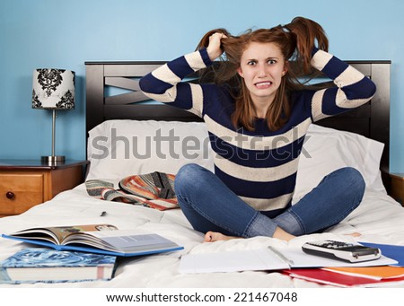 Attractive teen sitting on her bed, surrounded by her school books pulling on her hair in frustration.