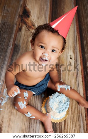 Cake Smash!  Adorable baby boy sitting on the floor, looking up, and enjoying a birthday cake.