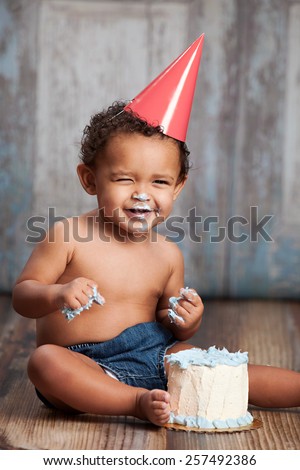 Adorable baby boy, sitting on a wood floor, wearing a party hat and eating a small cake.