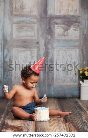 Adorable baby boy, sitting on a wood floor, wearing a party hat and eating a small cake.  Room for your text.