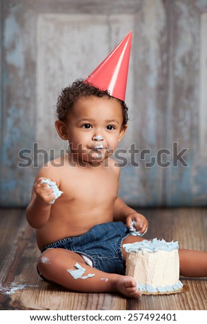 Adorable baby boy, sitting on a wood floor, wearing a party hat and eating a small cake.
