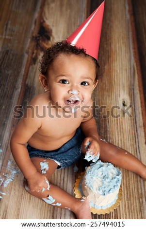 Adorable little boy sitting on a wood floor looking up from a cake he has been eating.