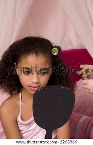 Adorable mulatto girl at a slumber party looking in the mirror as her friend puts her hair up in curlers.