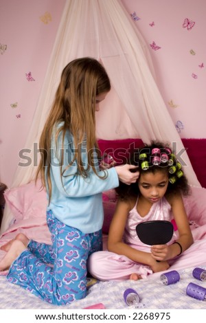 Adorable mulatto girl at a slumber party looking in the mirror as her friend puts her hair up in curlers.